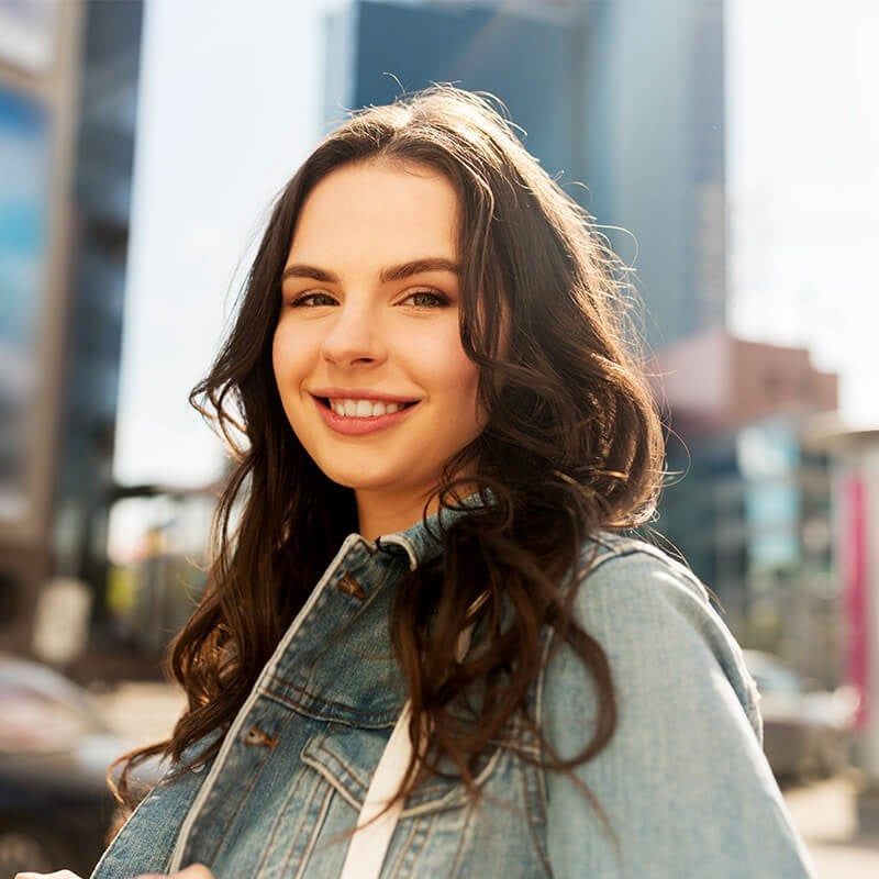 A smiling person with long dark wavy hair, wearing a denim jacket, outdoors in an urban setting with blurred buildings.