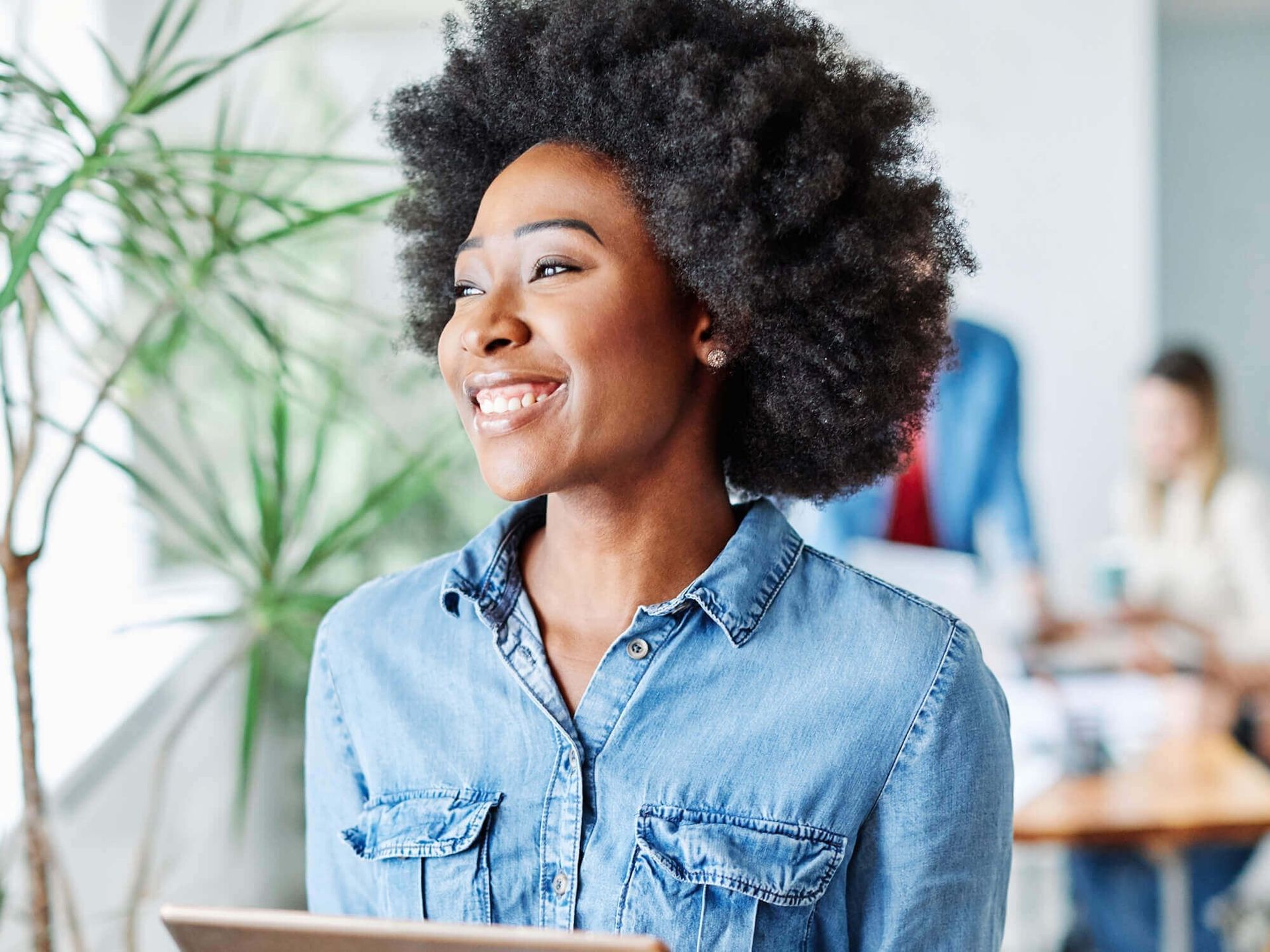 A smiling person in a denim shirt holds a tablet in a bright office space with a plant in the foreground.