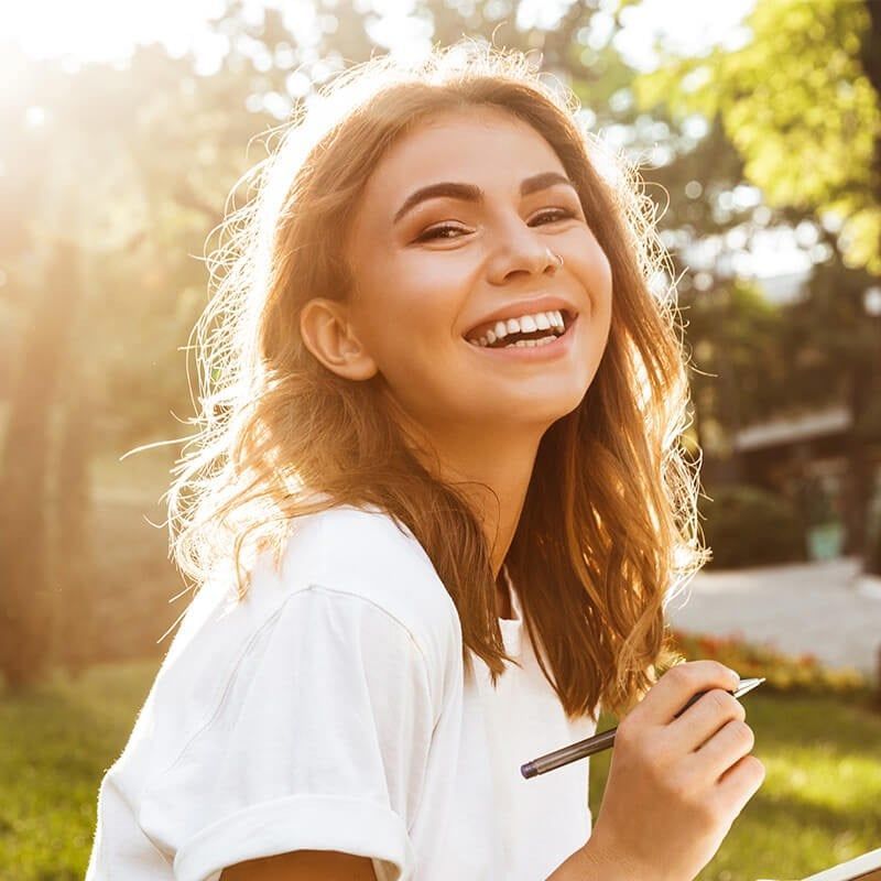 A smiling person holding a pen in a sunlit park, with soft, golden-hour light highlighting their hair.