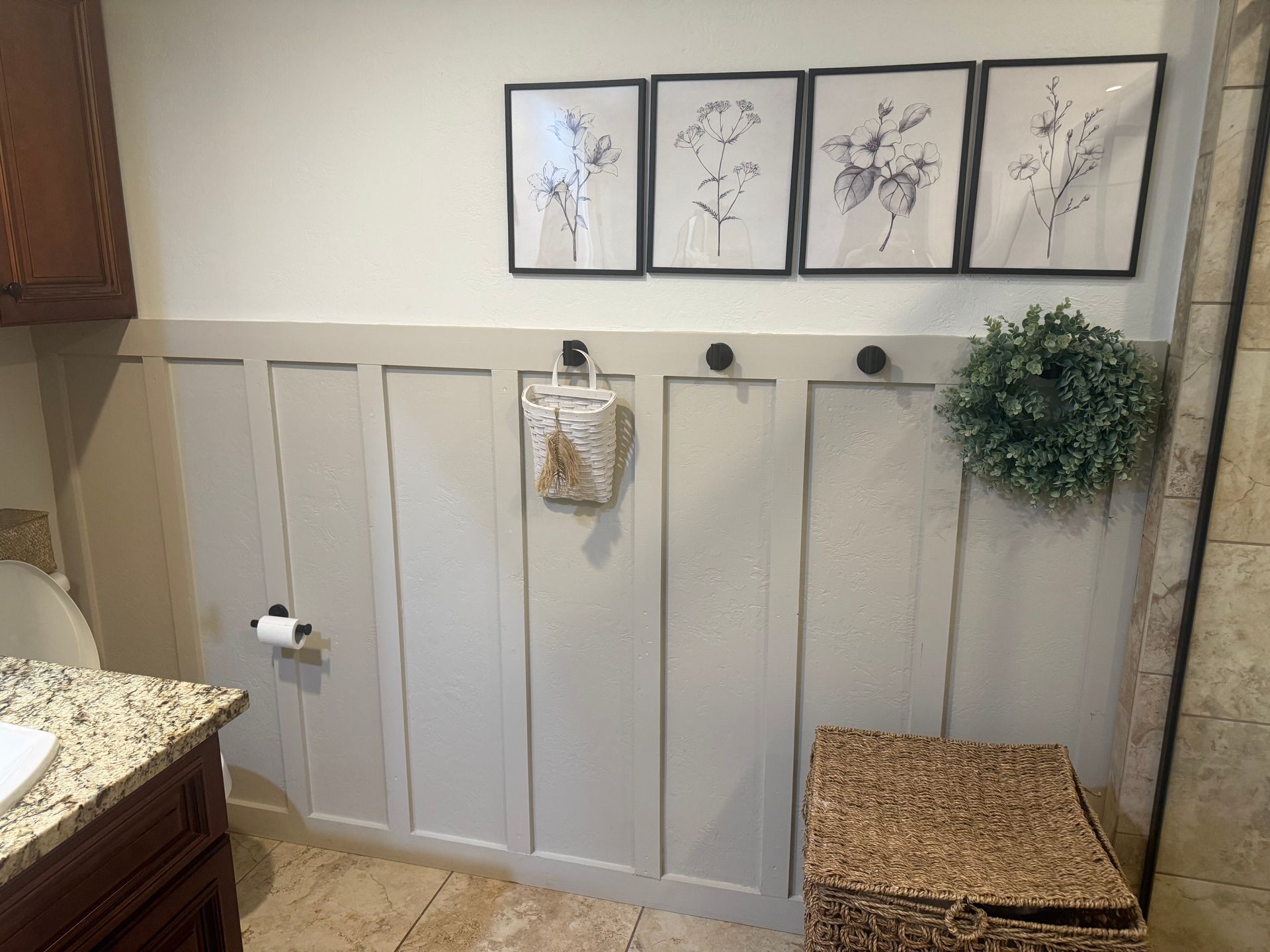 Light gray wainscoted wall with framed art, hooks, a hanging towel, and a wicker bench with plant.