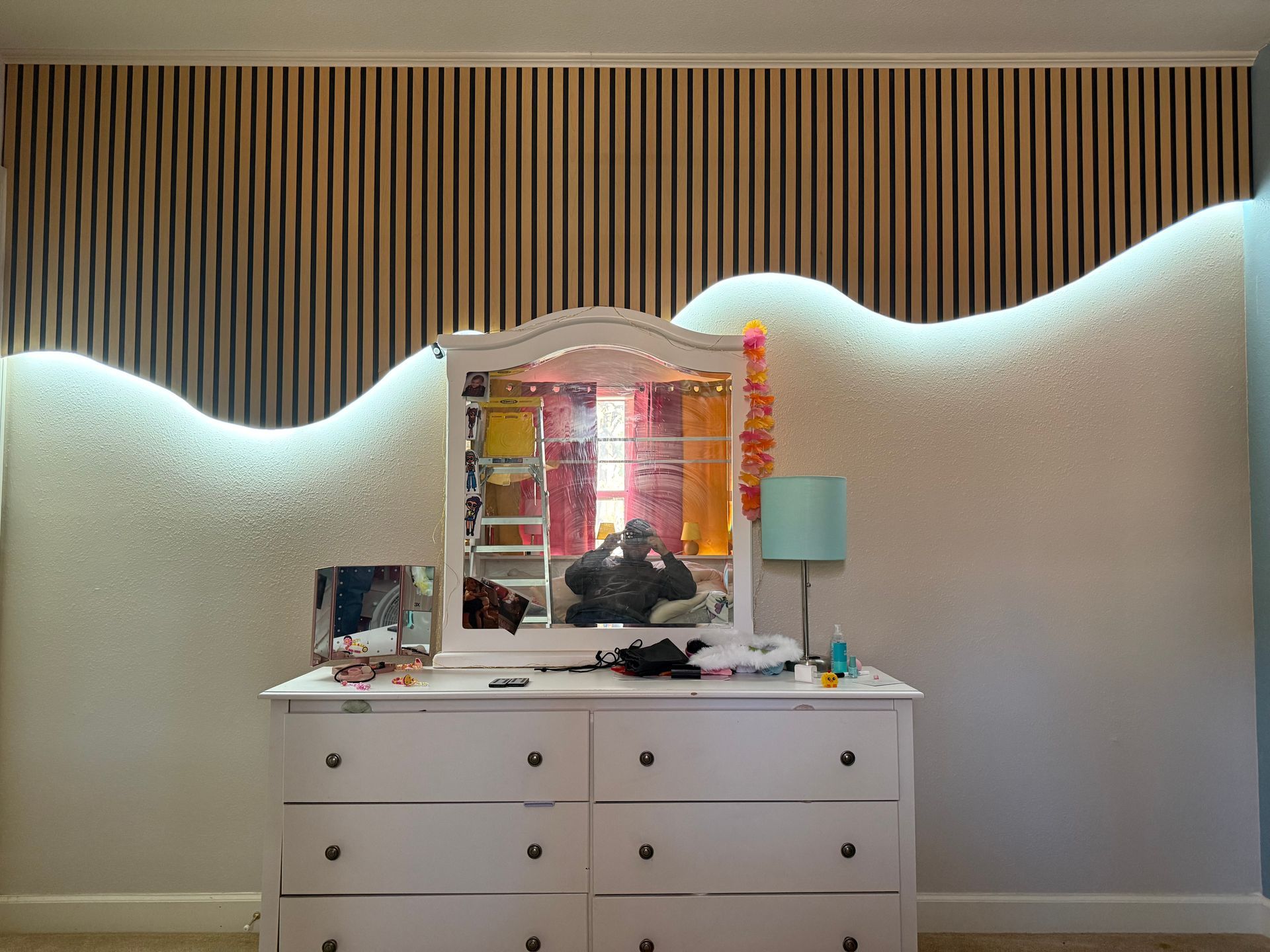 White dresser with mirror in a bedroom, decorated with small items and a wavy white-and-wood accent wall behind it.