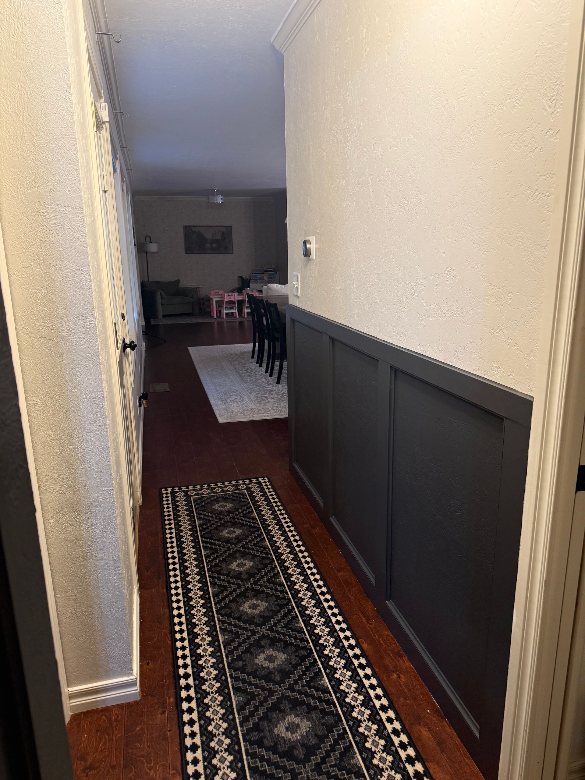Narrow hallway with dark wainscoting, runner rugs, and a view into a living room at the end.