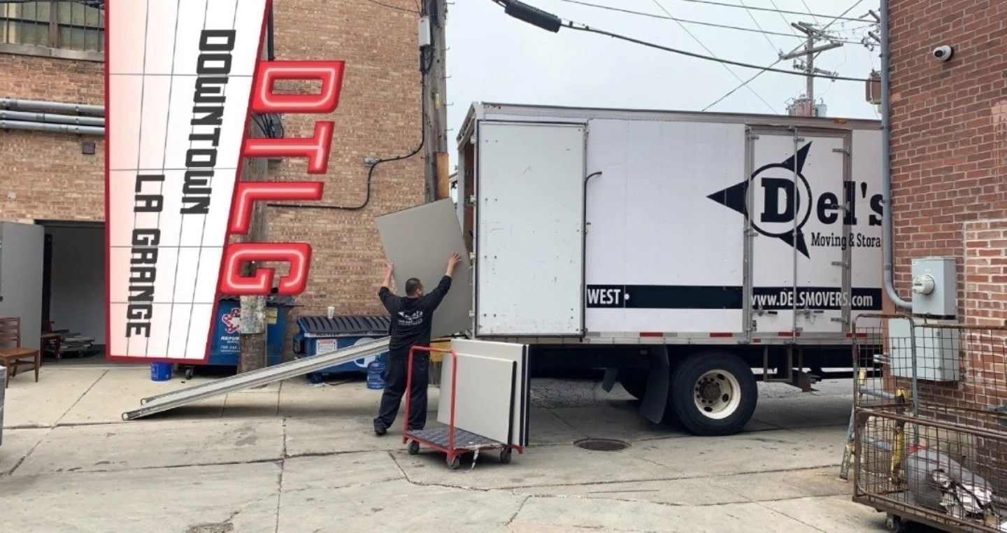 Man loading a moving truck with white panels in a downtown alley. Truck is marked with “Del’s”. Sign in the background: