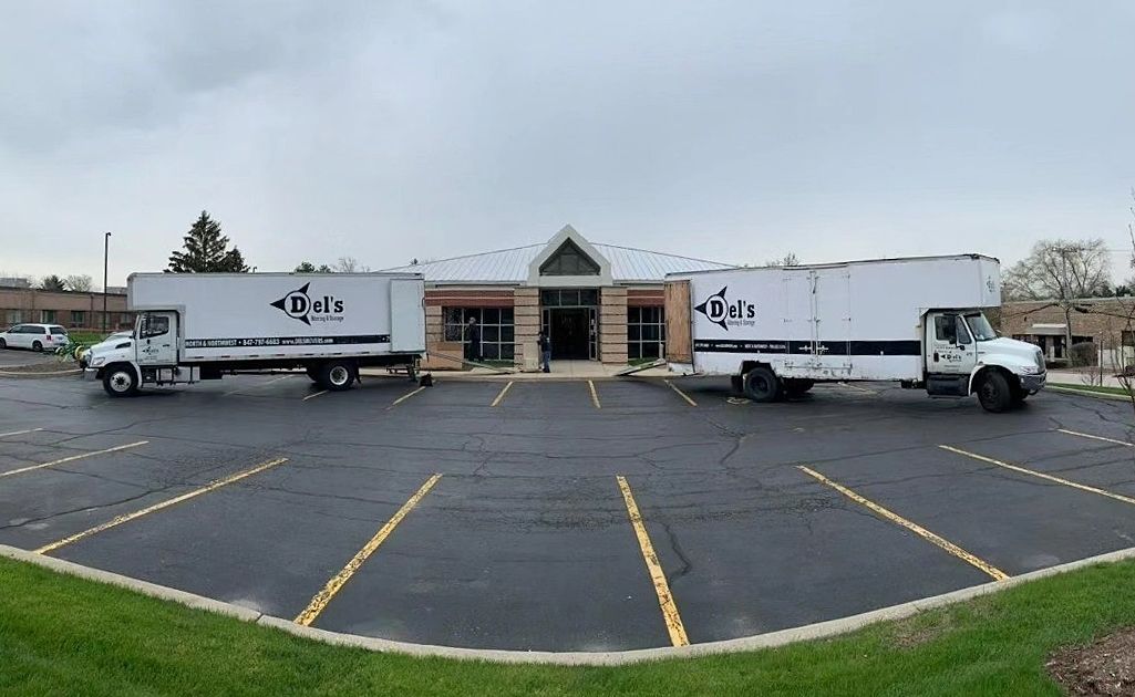 Two white moving trucks parked in front of a building with a light-colored roof on an overcast day.