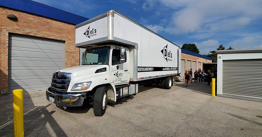 White moving truck parked in front of brick building with open garage doors and people near them.