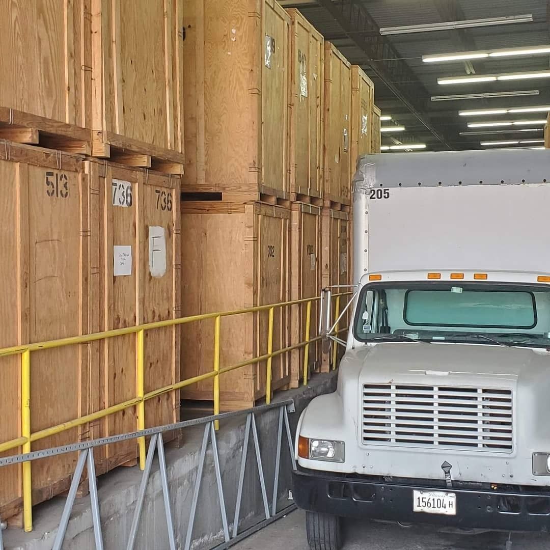 A white truck loading cargo containers at a storage facility; rows of large wooden crates.