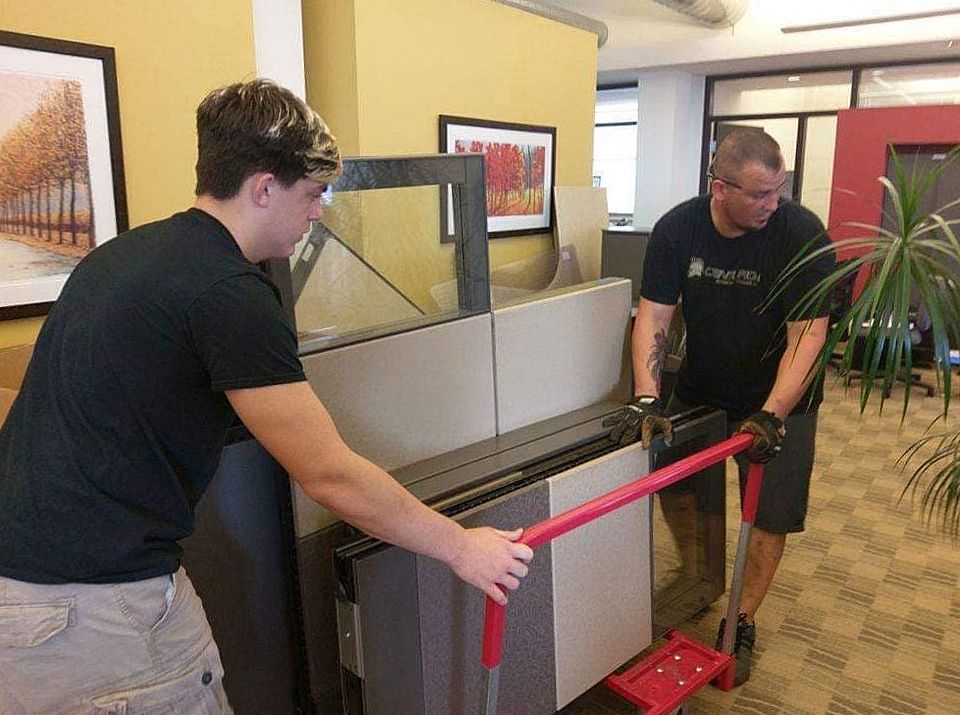 Two men moving office furniture with a hand truck in an office.