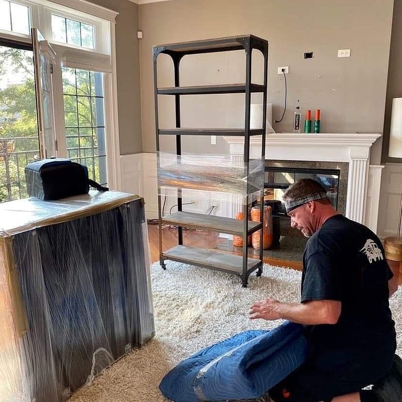 Man wrapping furniture, bookcase, in a living room with fireplace and window.