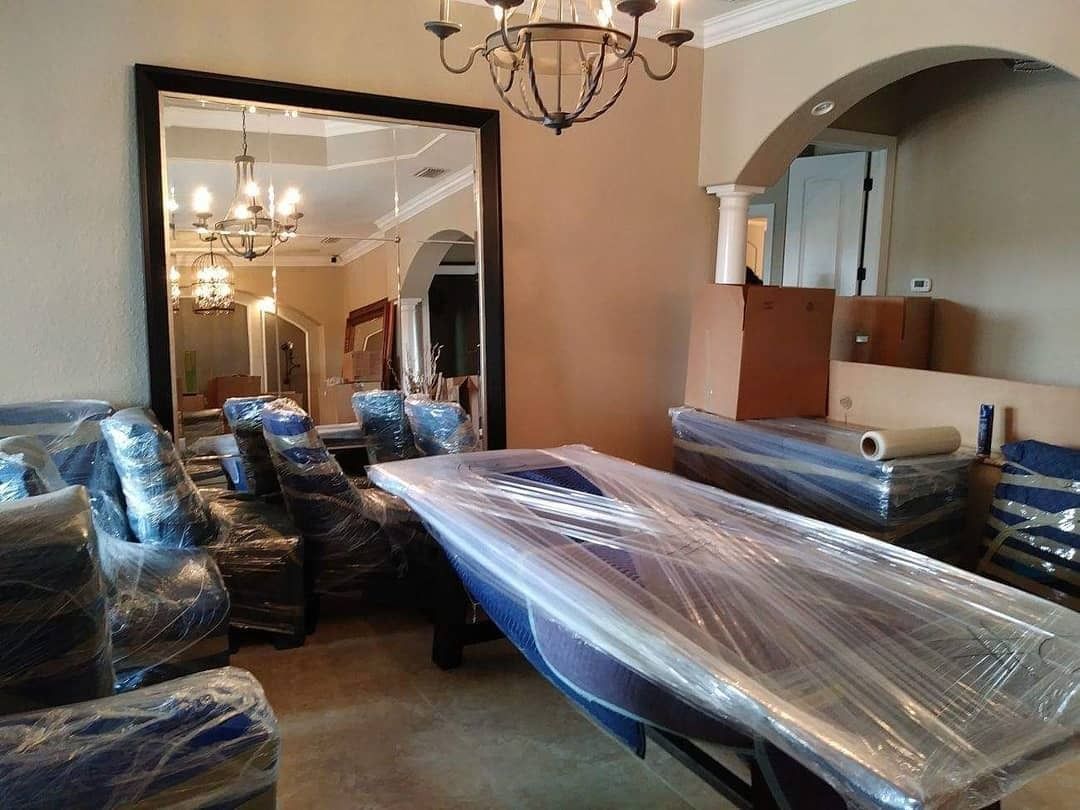 Furniture wrapped in plastic, ready for moving, in a room with a large mirror and chandelier.