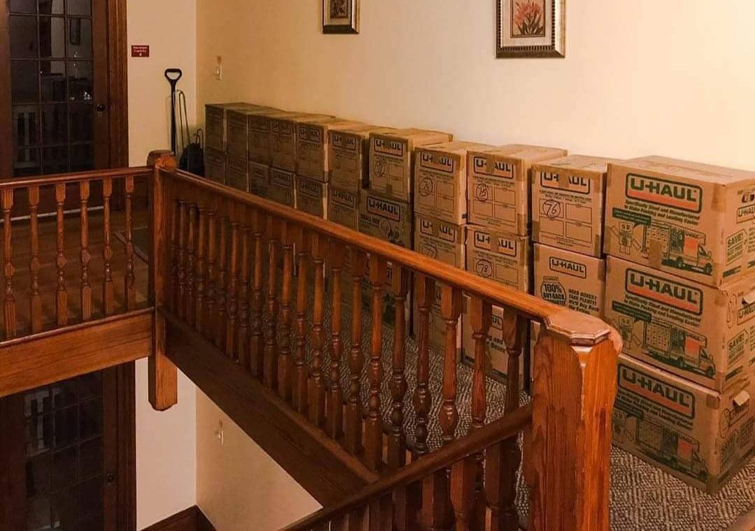 Boxes stacked on a landing above a wooden staircase. Brown boxes line the wall.