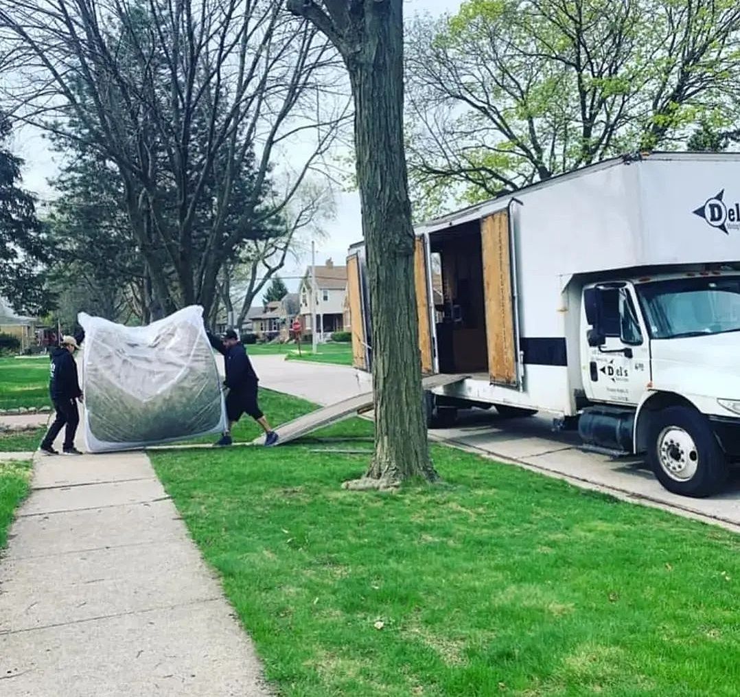Two people moving a large, covered object towards a white moving truck parked on a residential street.