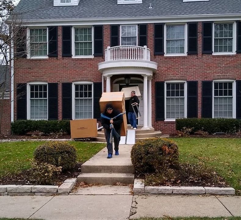 Two people carrying boxes and items from a brick house with a white porch.