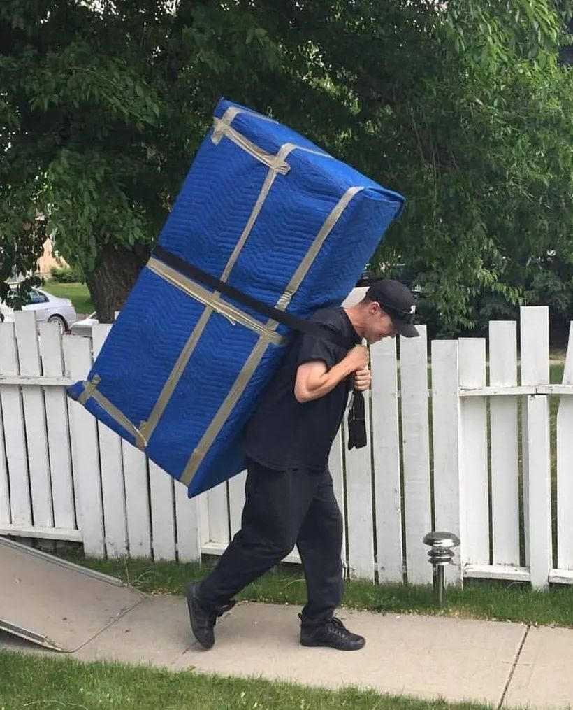 Man carrying a large, blue-padded, wrapped object on his back while walking on a sidewalk near a white fence.