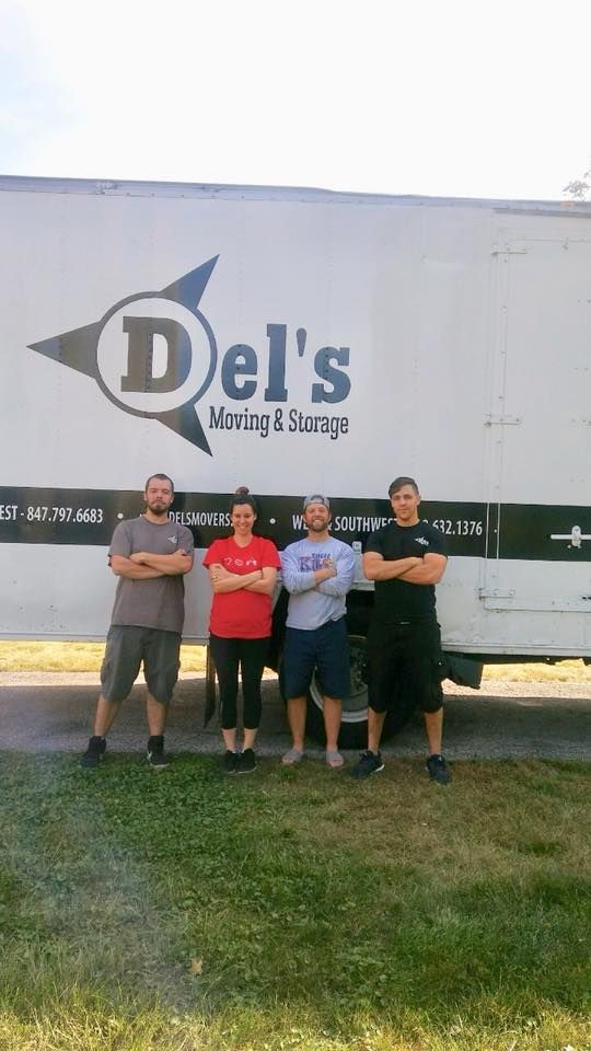 Four people posing in front of a Del’s truck logo, outdoors on grass