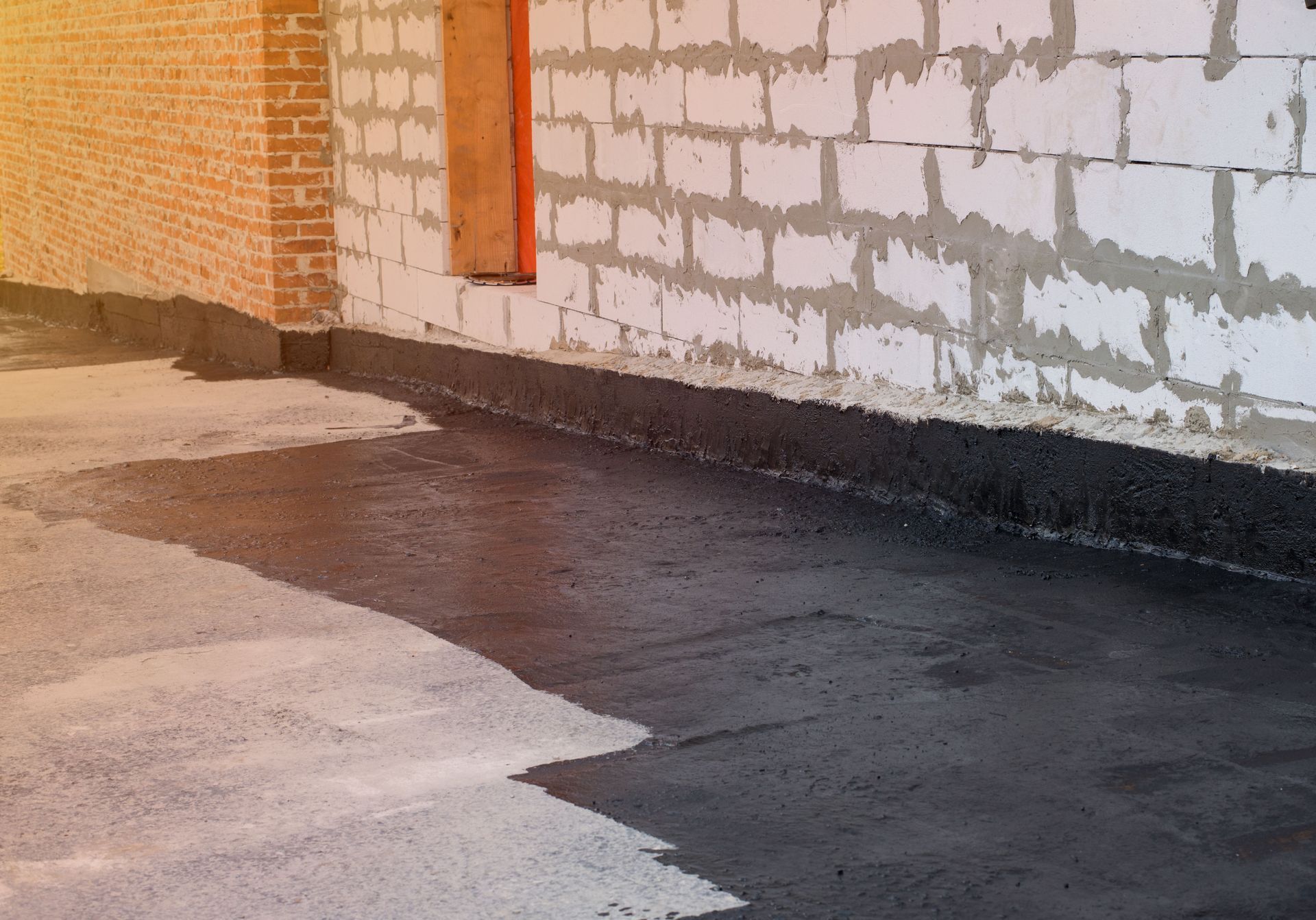 Black waterproof bitumen coating applied to a concrete floor and the base of a white block wall.