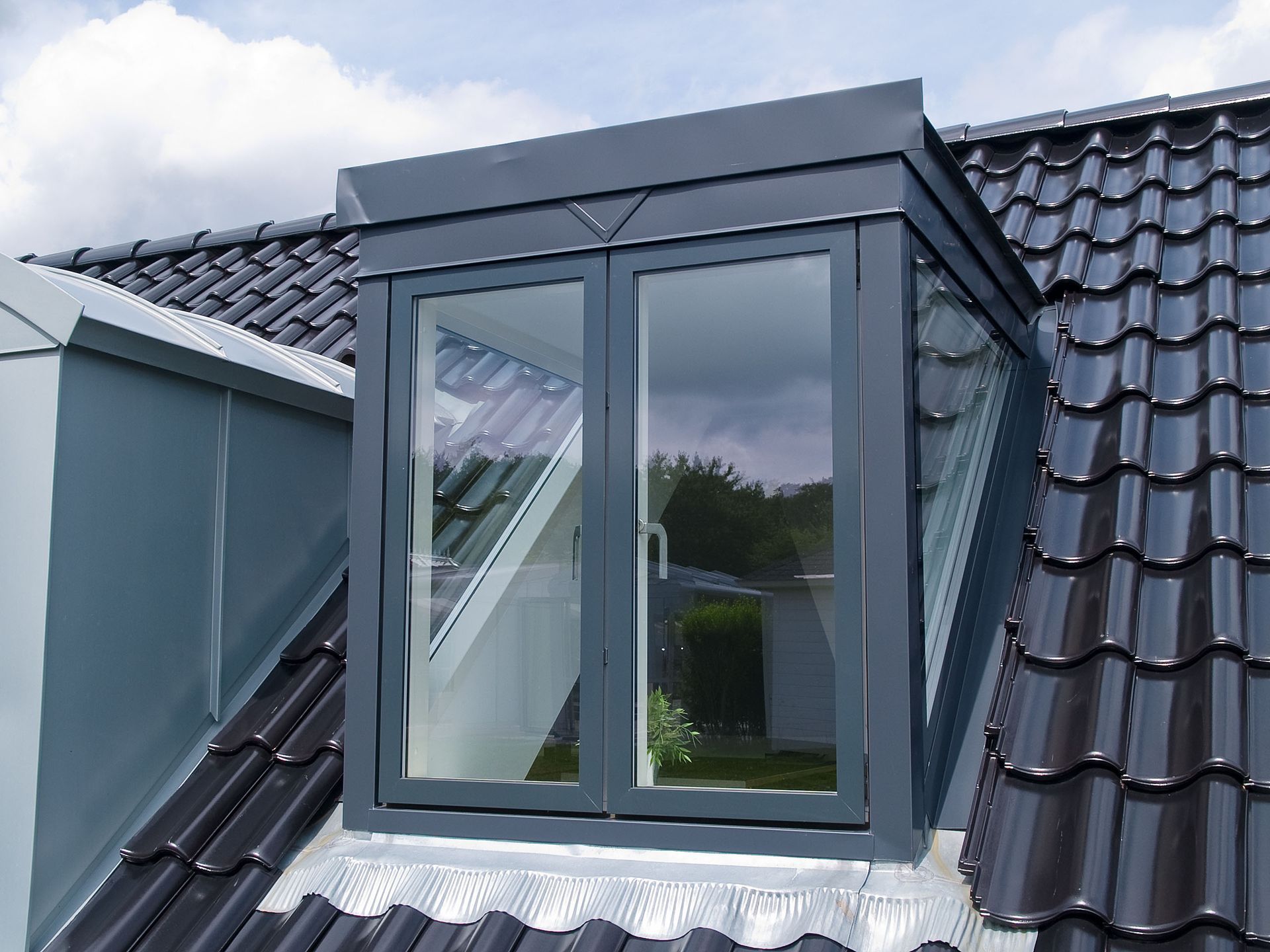 A modern, dark grey dormer window set into a dark tiled sloped roof under a cloudy sky.