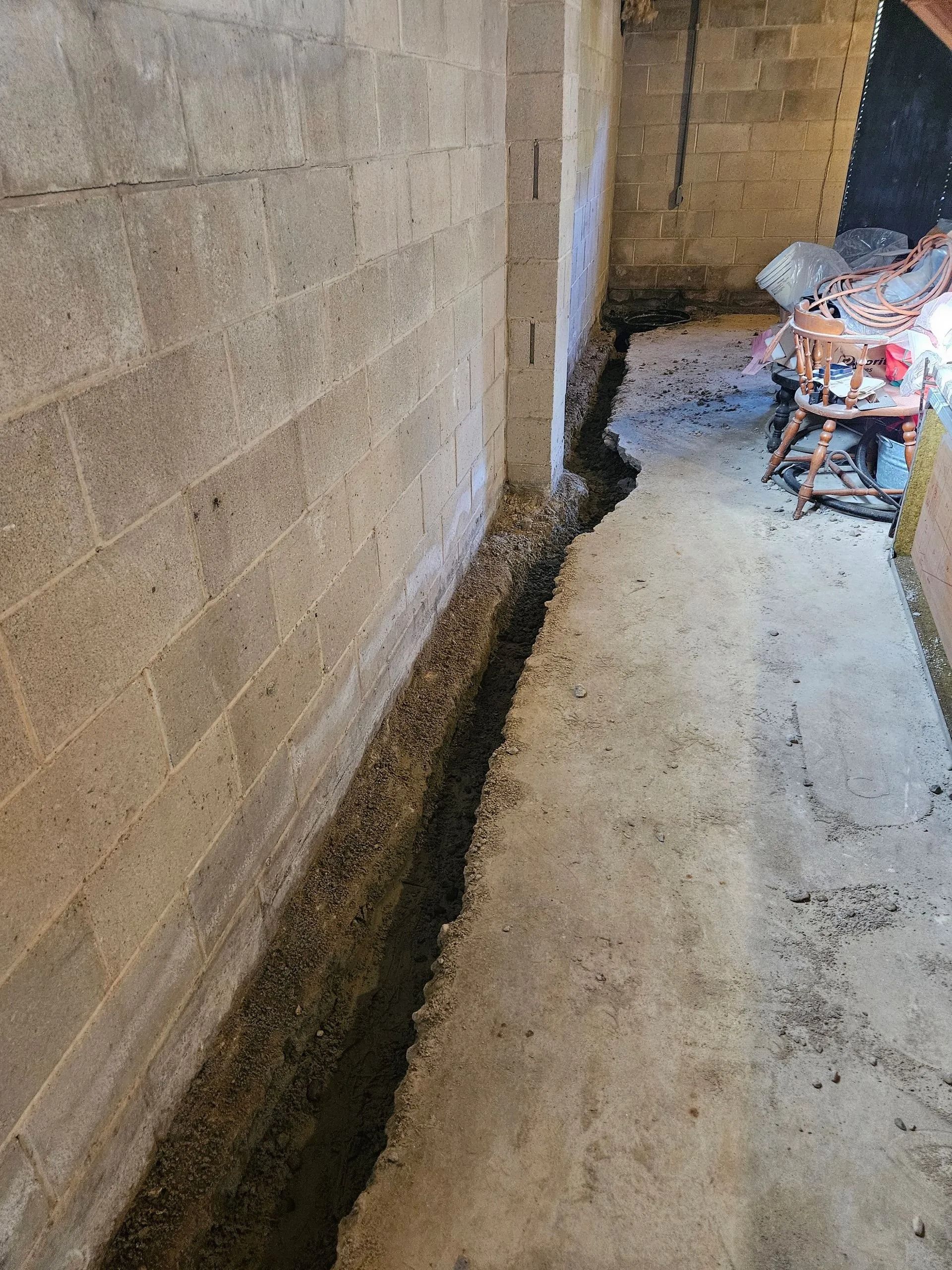 A trench cut into a concrete basement floor along a cinder block wall, preparing for a drainage system installation.