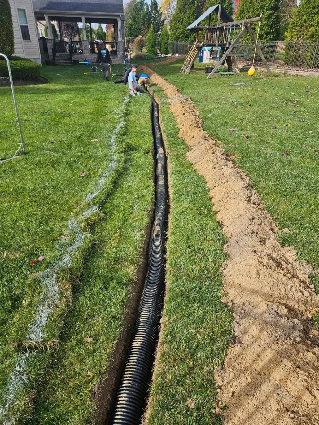 A black corrugated drainage pipe lies in a trench dug through a grassy backyard, next to a pile of excavated soil.