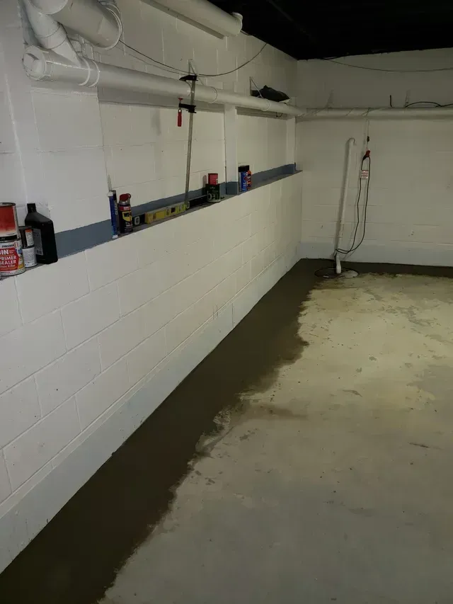 A flooded basement corner with water pooled along the base of white concrete block walls under exposed ceiling pipes.
