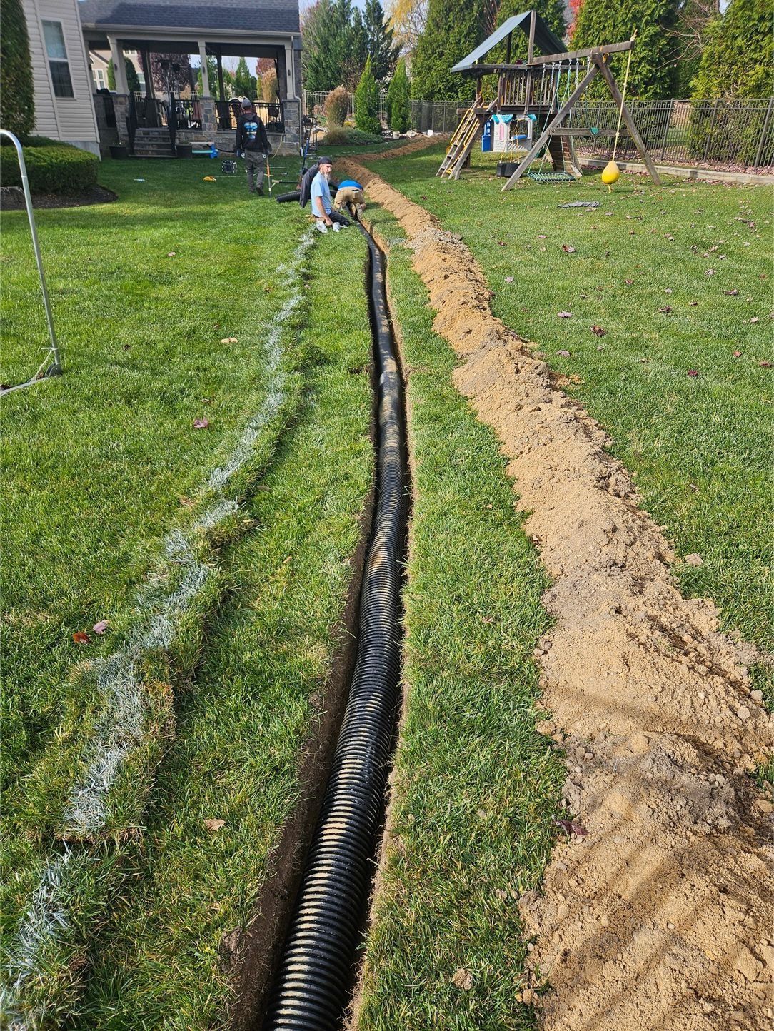 A black corrugated drainage pipe sits in a trench dug through a grassy yard, with piles of displaced dirt beside it.