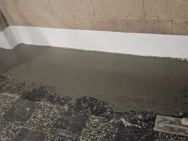 Freshly applied gray concrete patch on a floor, bordering a wall with a painted white baseboard.