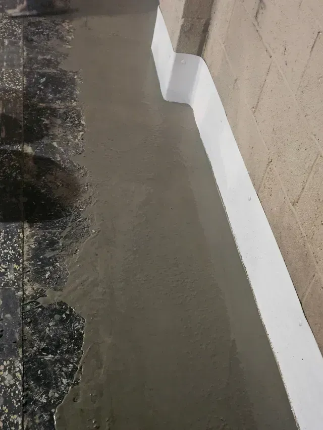 A thick layer of wet, grey concrete is being applied along a wall with a white waterproof membrane base.