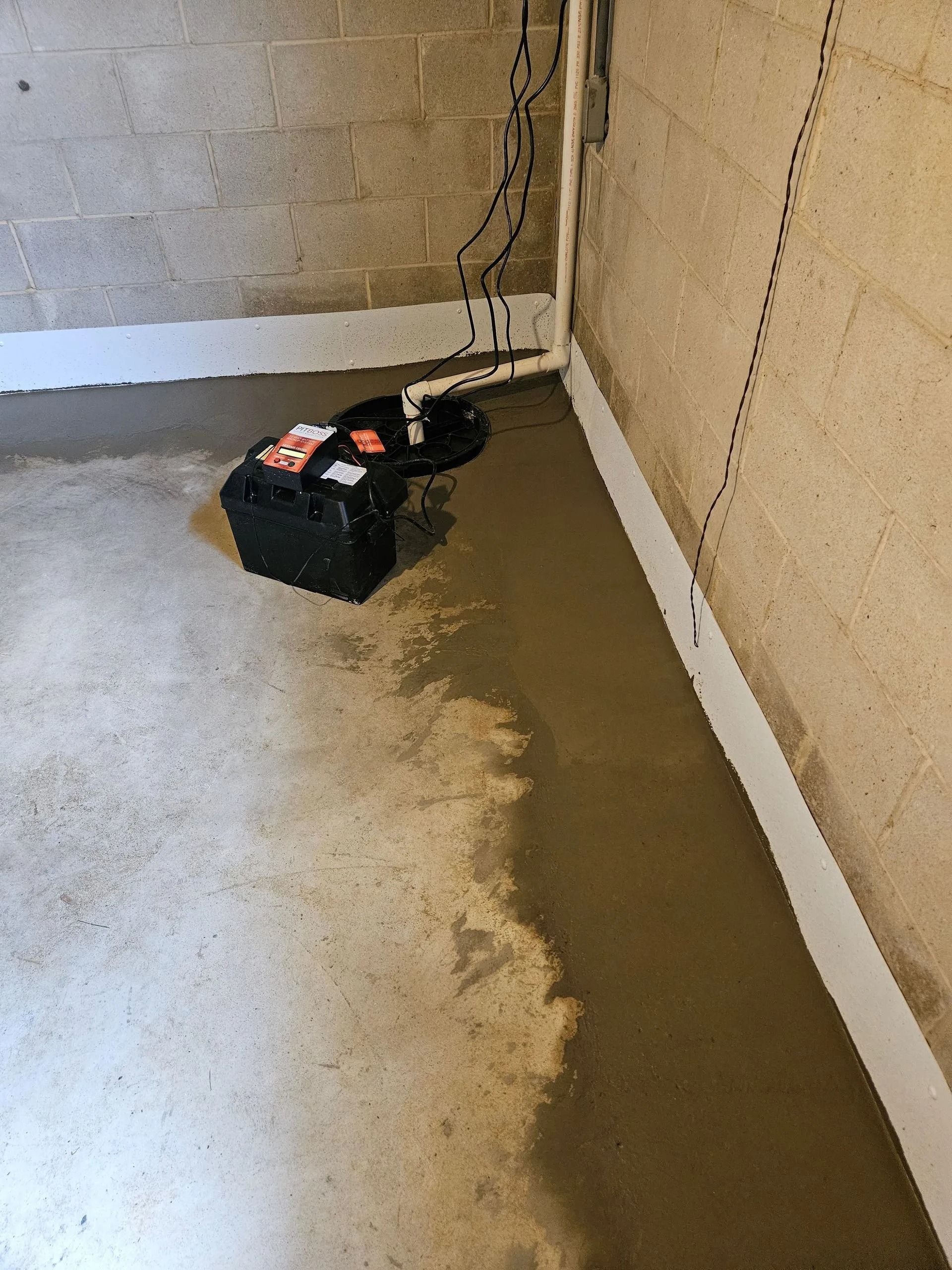 A basement corner with a sump pump, battery backup, and a damp concrete floor near the wall.