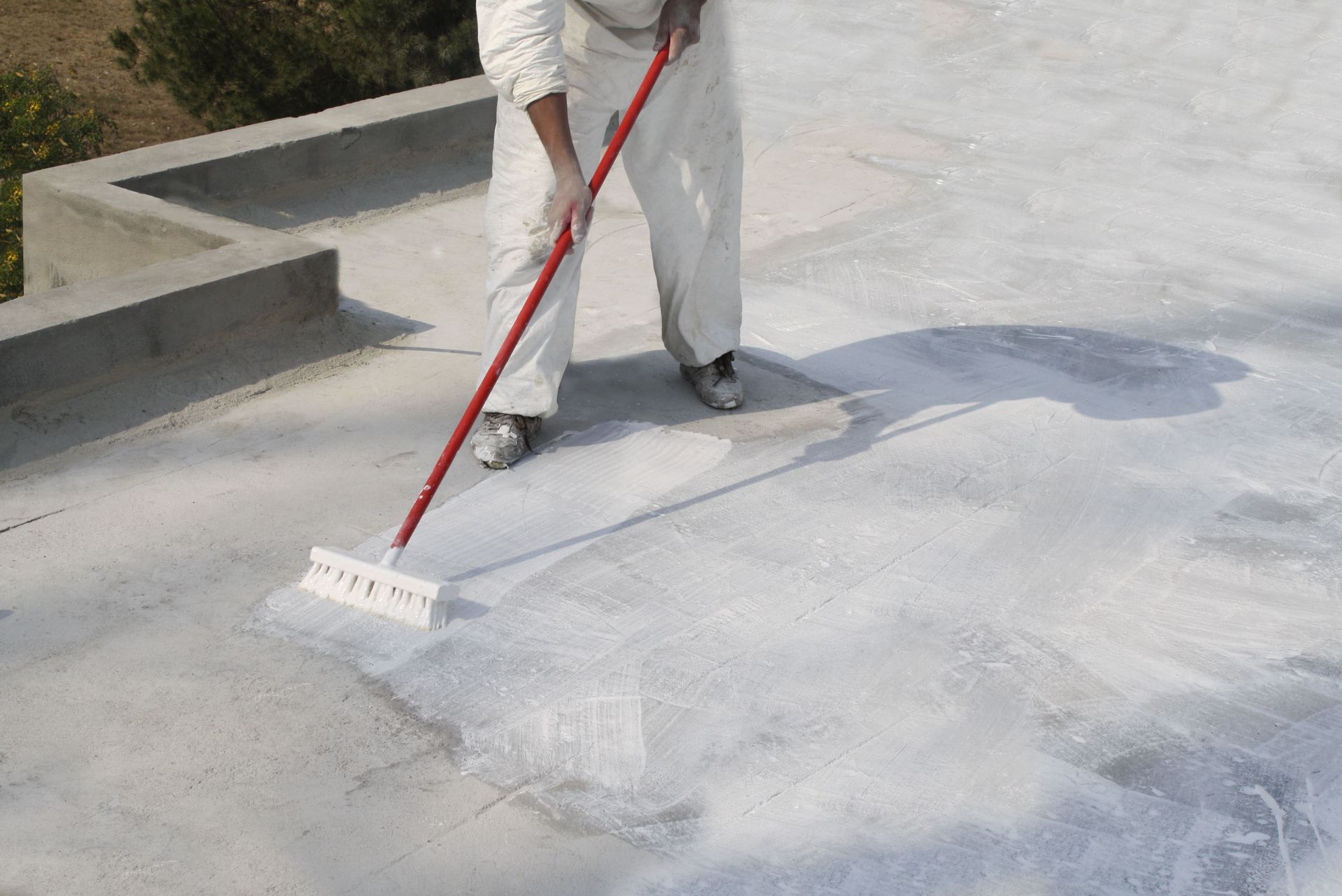 A person in white protective gear applying white sealant to a concrete roof surface with a long-handled brush.