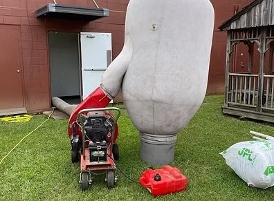 Large gray inflatable figure tethered to a red blower on a lawn beside a building, with sandbags nearby