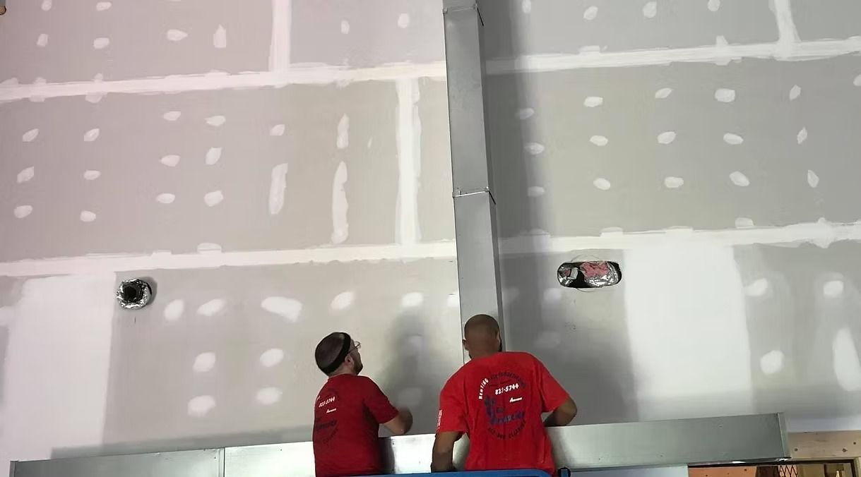 Two workers in red shirts installing drywall on a partially finished wall with taped seams and patch marks.