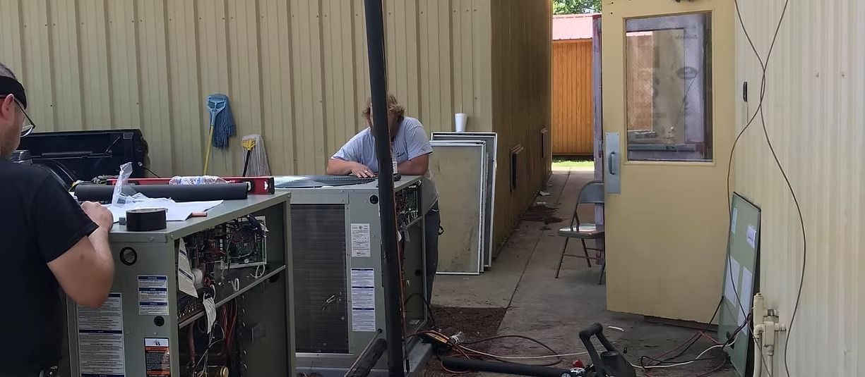 Two workers operate equipment beside a tan industrial building with an open door and cables on the ground