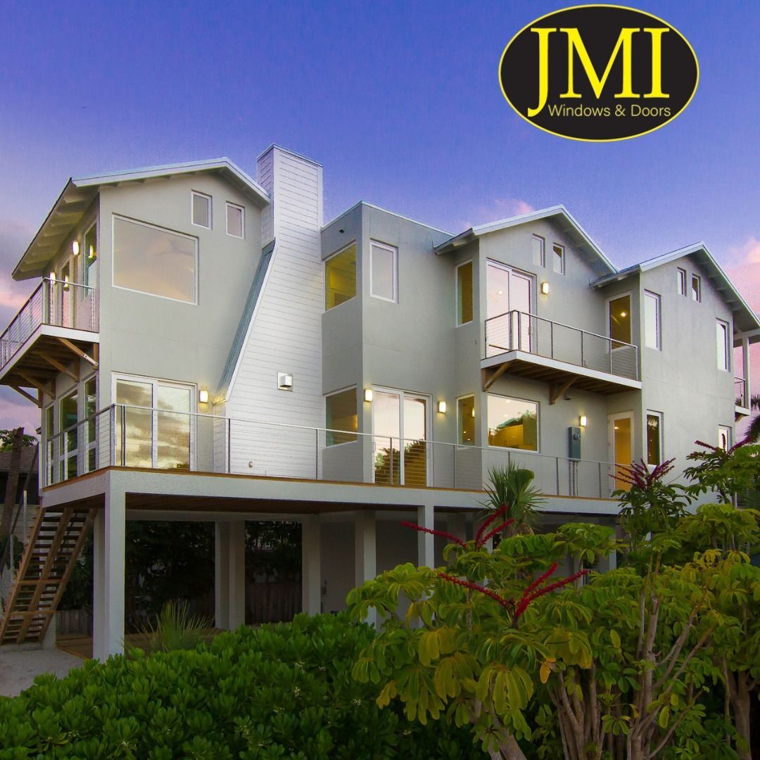 A modern three-story house on stilts at dusk, featuring large glass windows, balconies, and a JMI Windows & Doors logo.