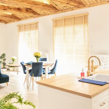 Bright kitchen and dining area with wooden ceiling, white blinds, round table, and sunlit island counter