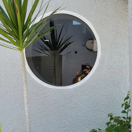 Round white-framed window in a textured wall, reflecting a person holding a phone and nearby green plants.
