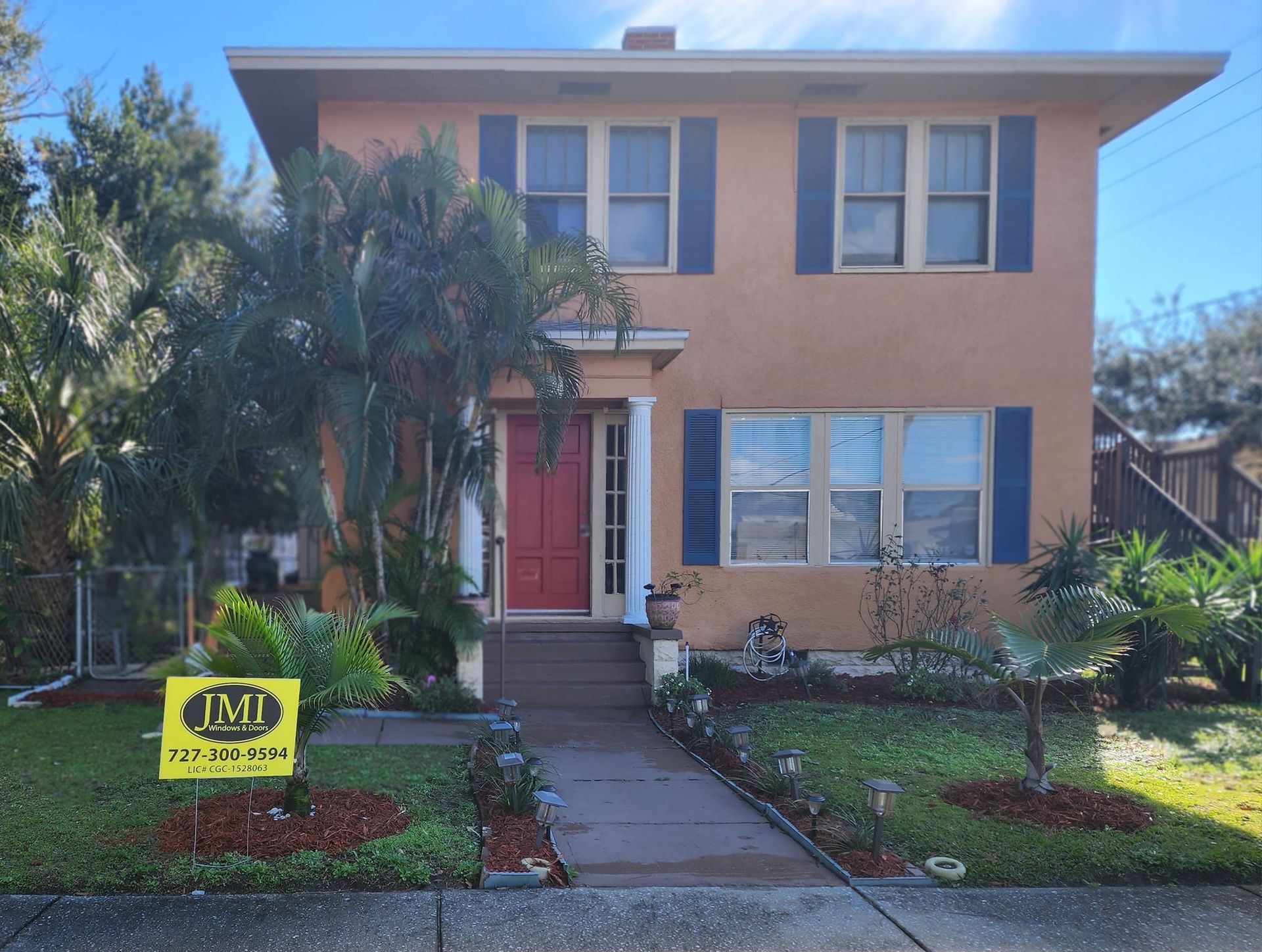 A two-story tan house with blue shutters, a red front door, a front porch, and a yellow real estate sign in the yard.