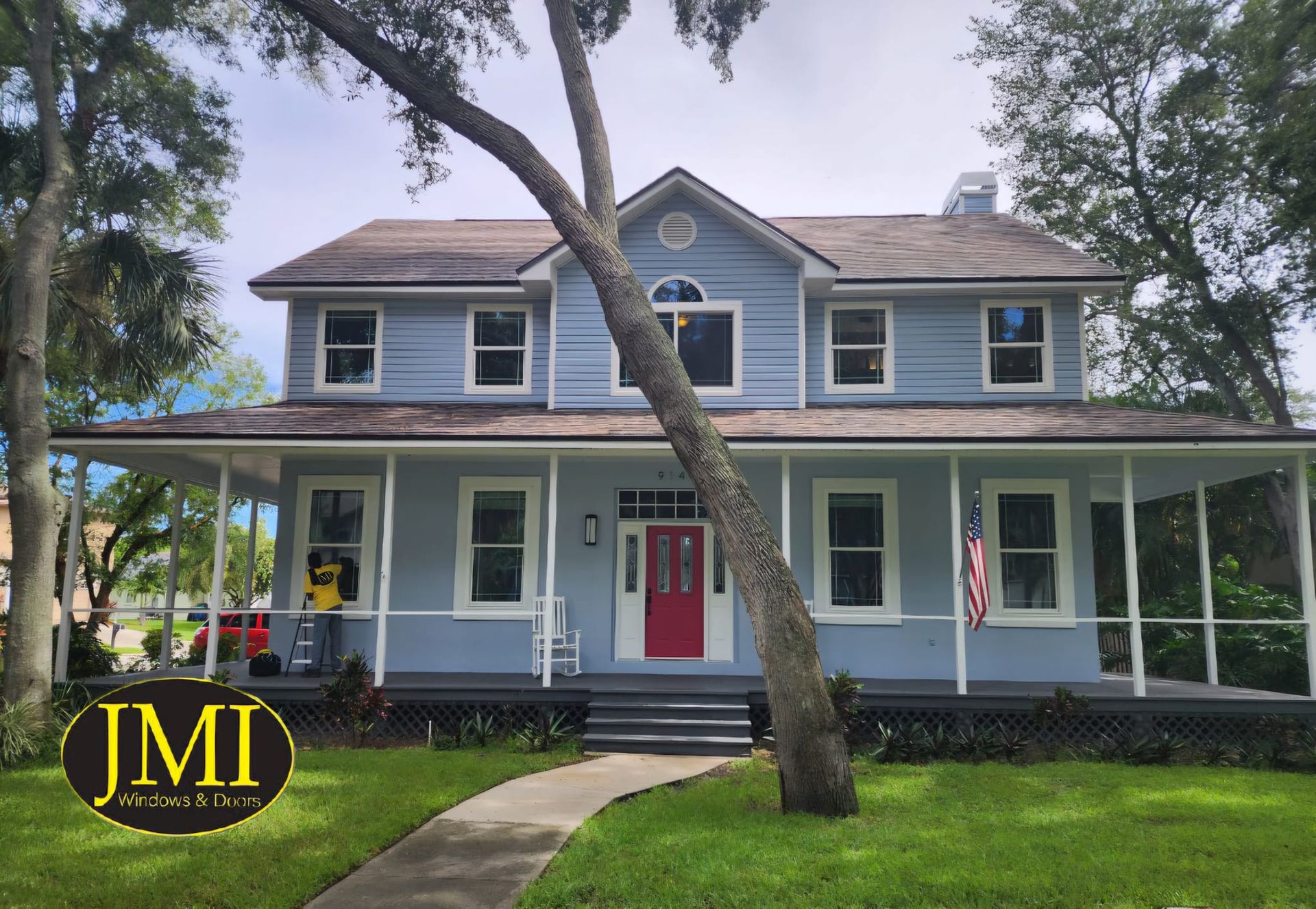 A two-story light blue house with a wrap-around porch, a red front door, and a tree in the front yard.