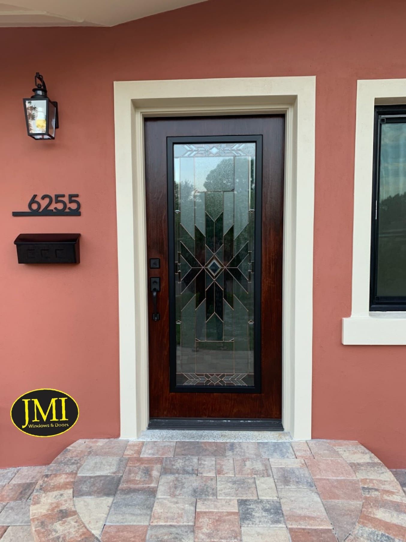 A dark wooden front door with a geometric frosted glass panel, flanked by house number 6255, a lamp, and a mailbox.