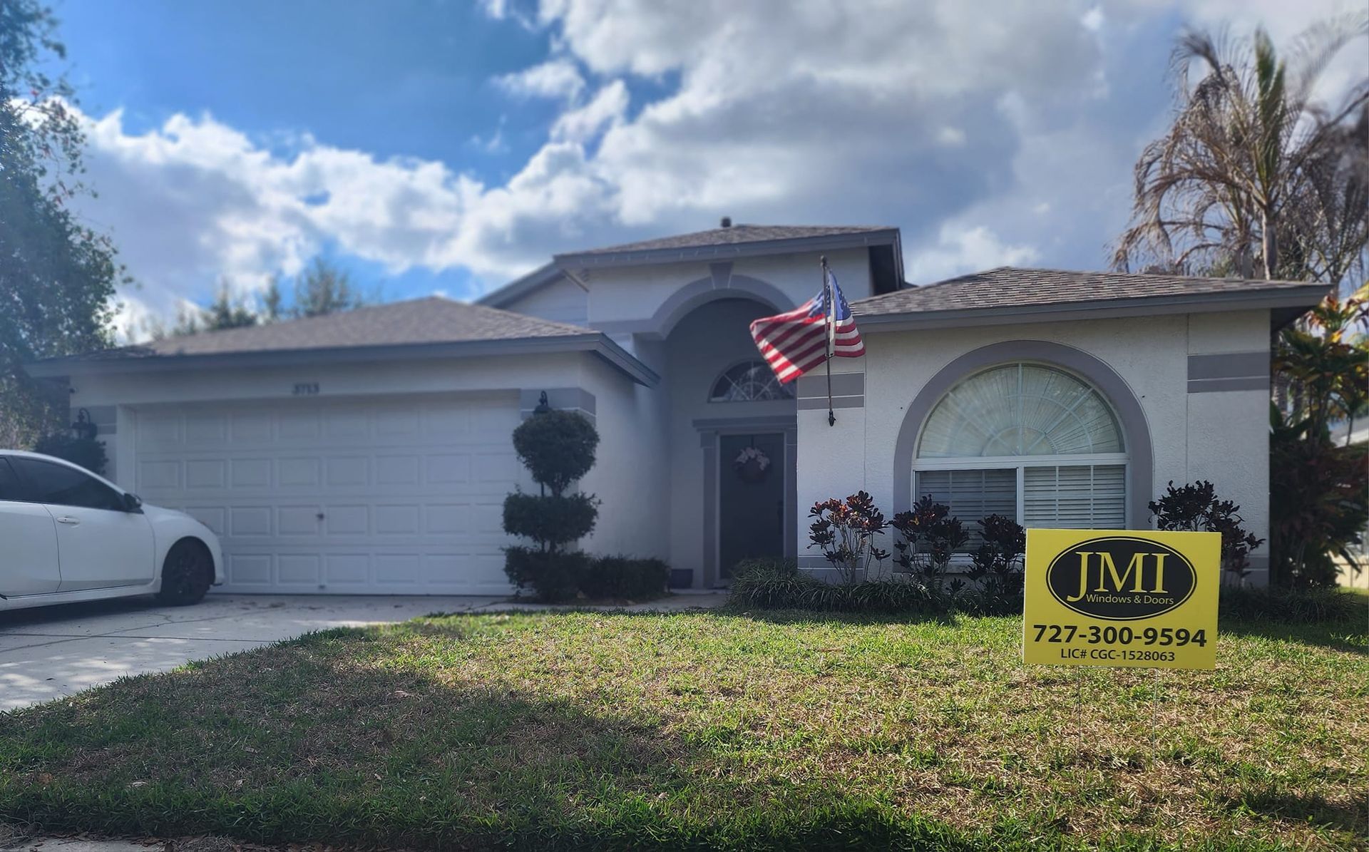 A single-story suburban house with white stucco, a two-car garage, an arched window, and a yellow sign in the front yard.
