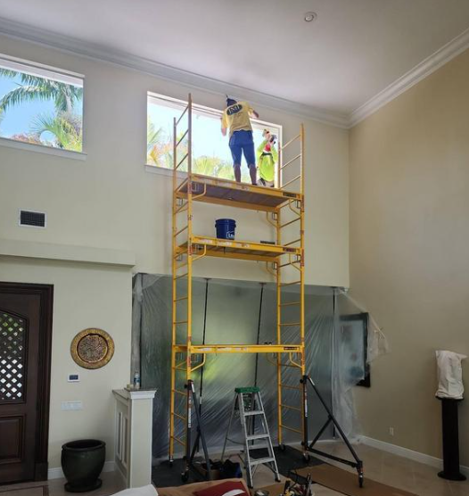 Worker on yellow scaffolding painting a high indoor wall in a living room.