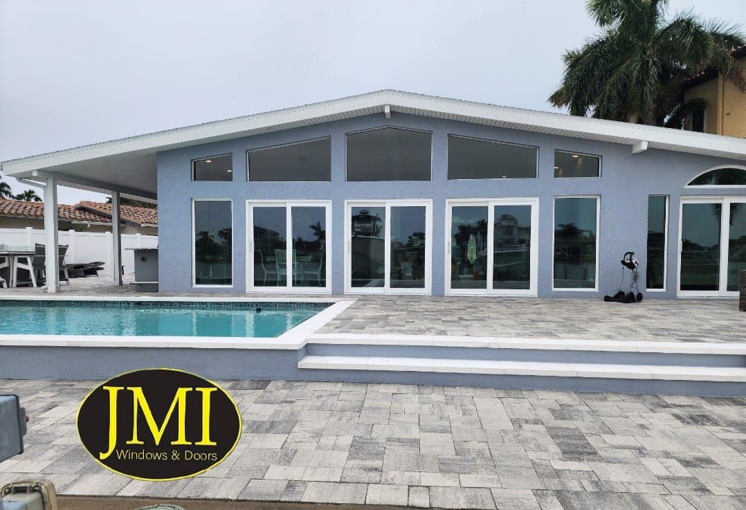 A light blue house with large sliding glass doors and windows, situated next to a swimming pool on a paved patio.