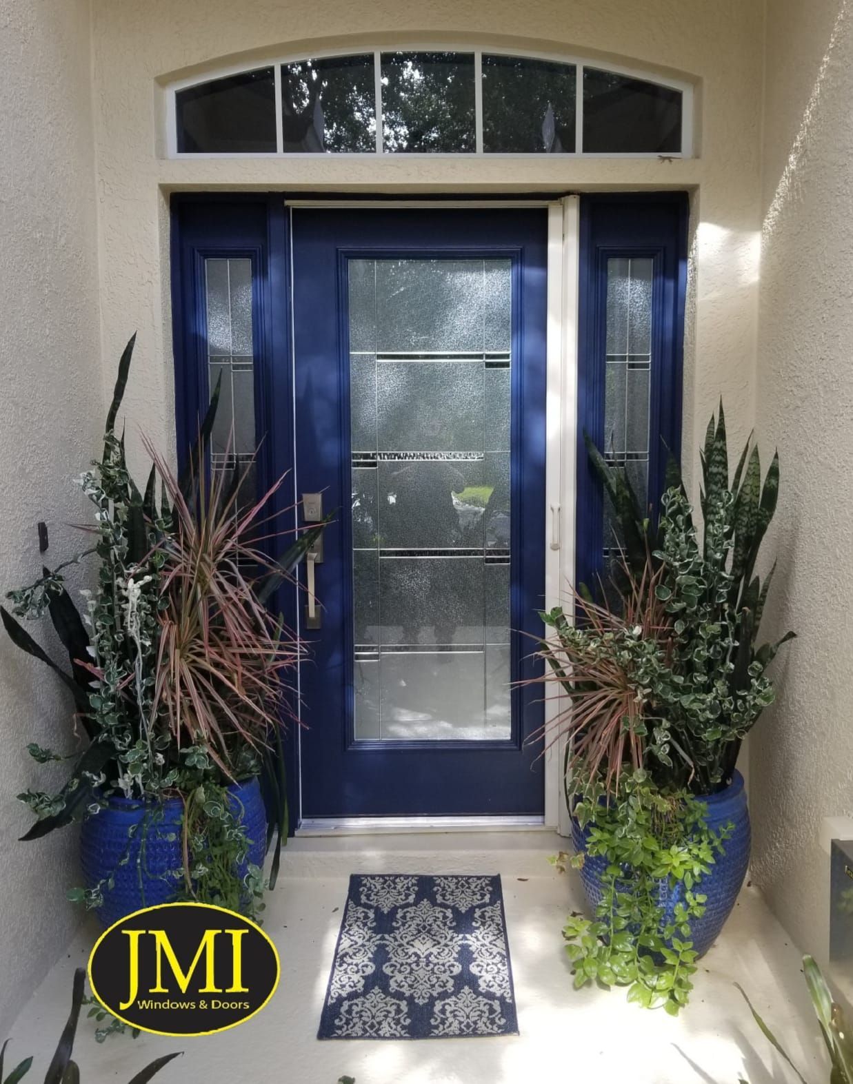 A blue front door with glass panels and a transom window, flanked by two large potted plants on a porch with a patterned rug.