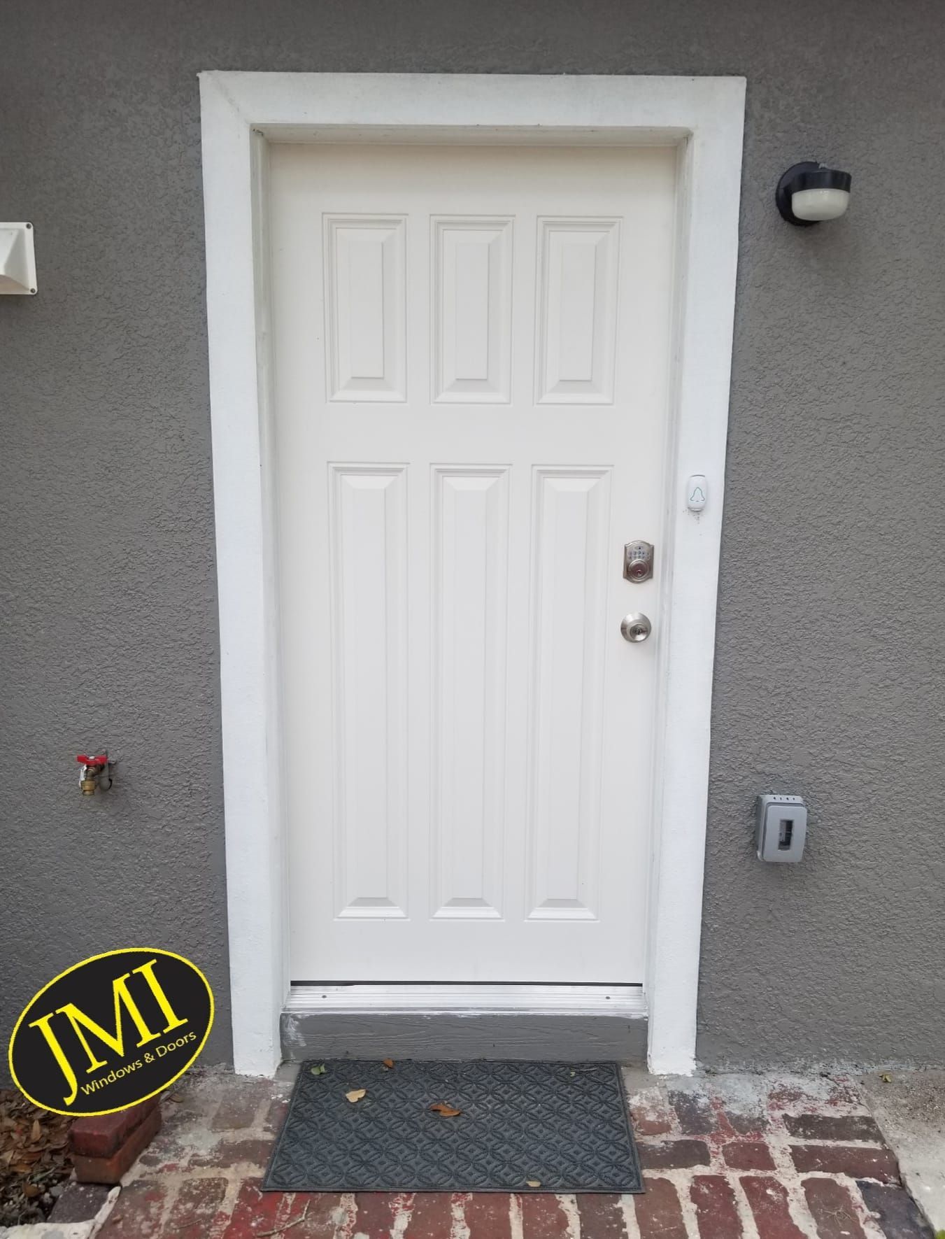A white six-panel door set in a gray stucco wall, featuring a black doormat and a small light above the entrance.
