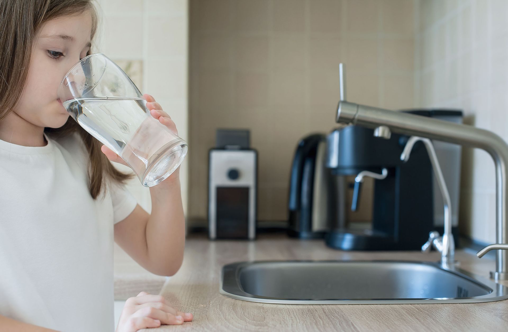 Girl drinking water from a glass in a kitchen next to a sink and water filter.