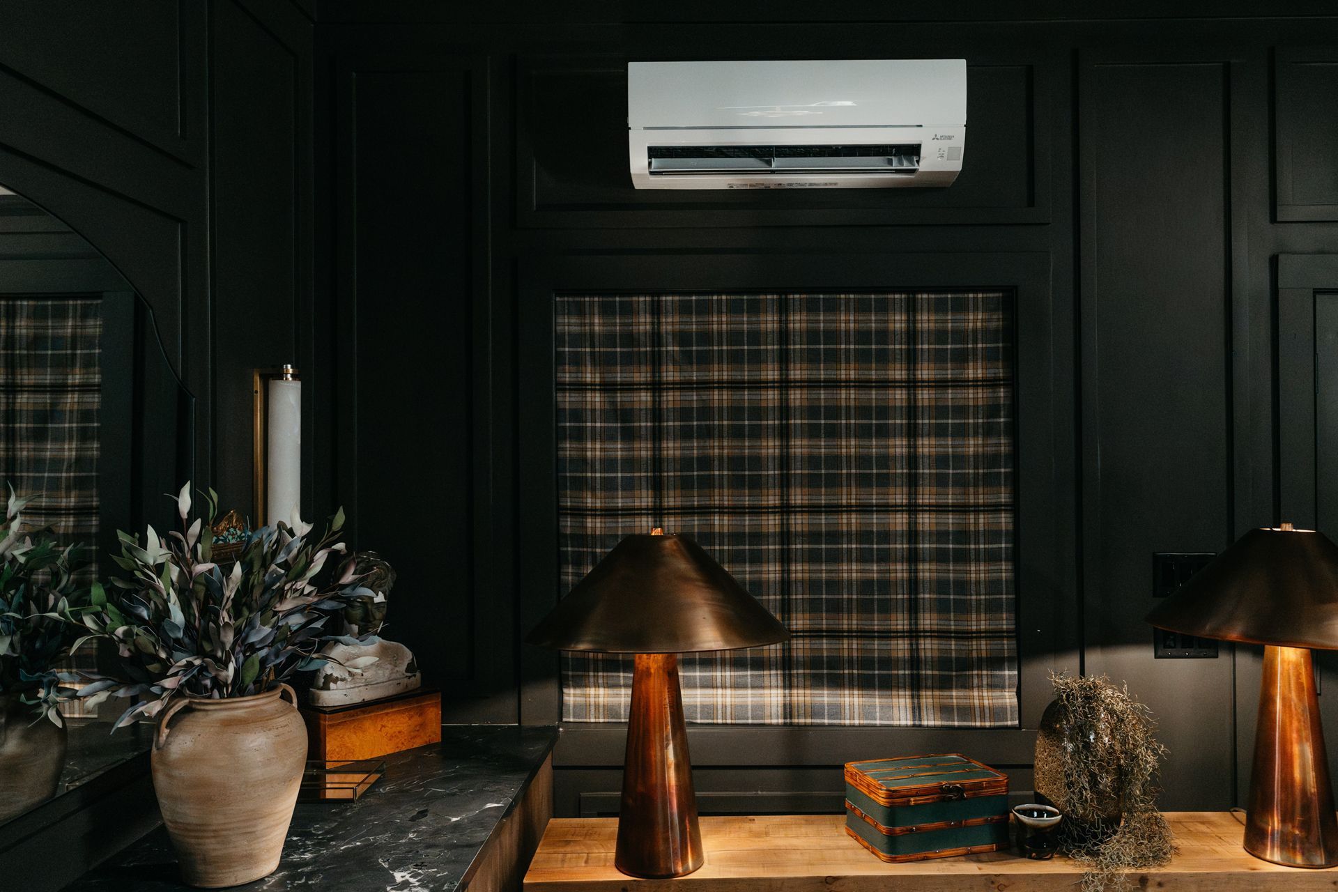 Dark paneled room with air conditioner, plaid curtains, lamps, and flowers on a table.