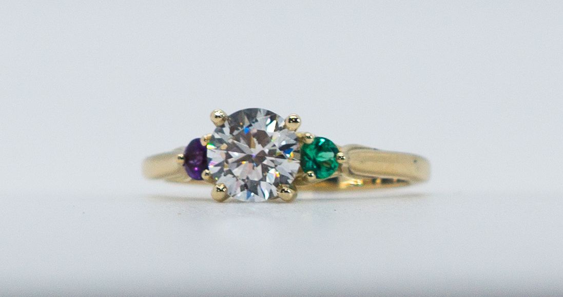 Gold ring with clear center stone and purple and green side gems on a light background