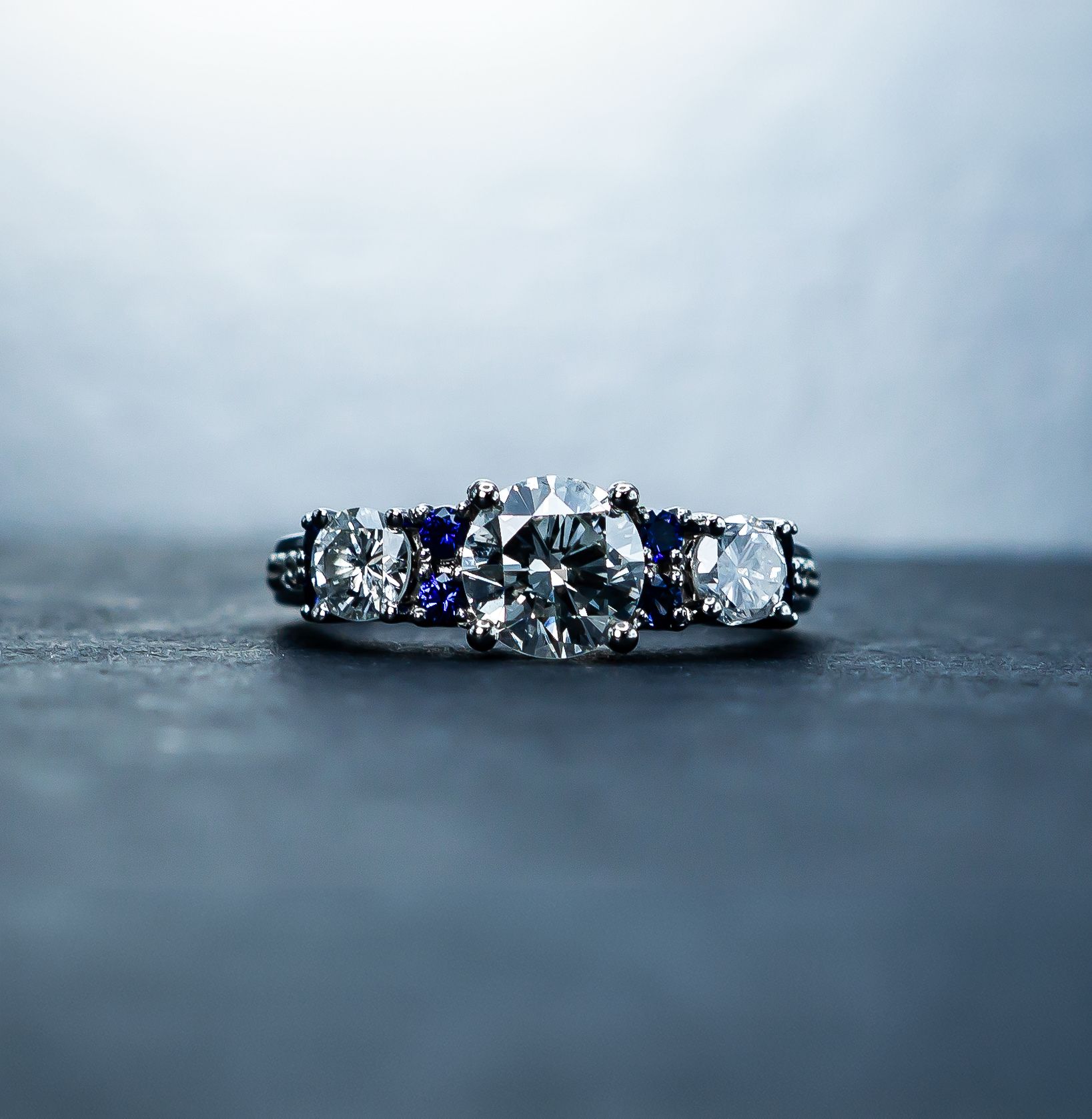 Diamond ring on a dark surface, with a large central stone and smaller blue accents in soft light
