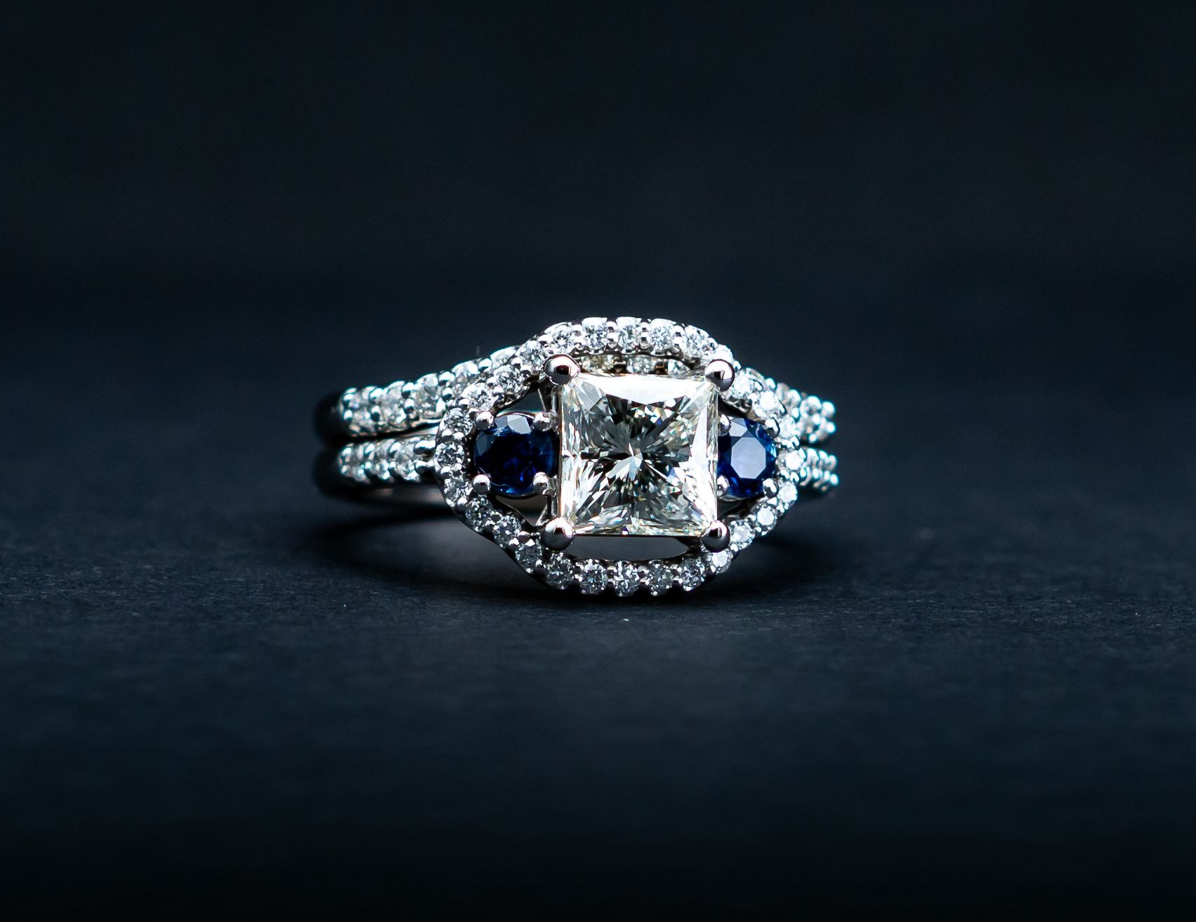 Diamond ring with a square center stone and blue side stones on a dark background