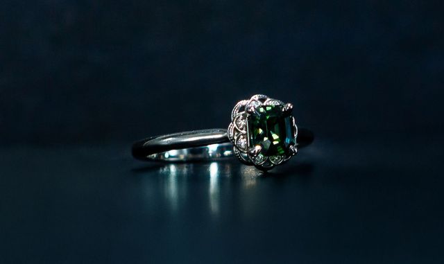 Dark gemstone engagement ring on a reflective black surface, lit against a dark background