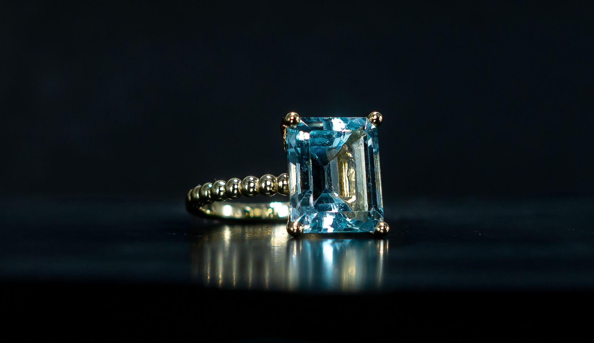 Blue gemstone ring with rectangular center stone on a dark reflective surface