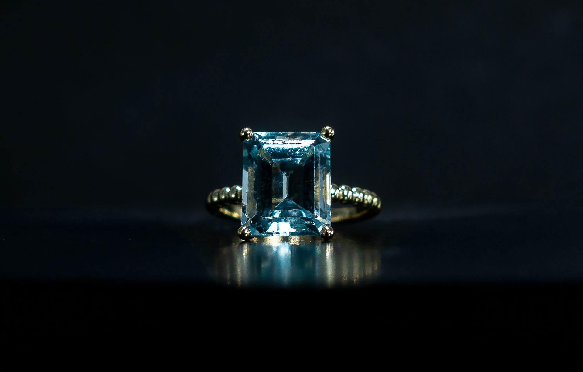 Blue gemstone ring on a dark reflective surface, with side diamonds and a glossy black background